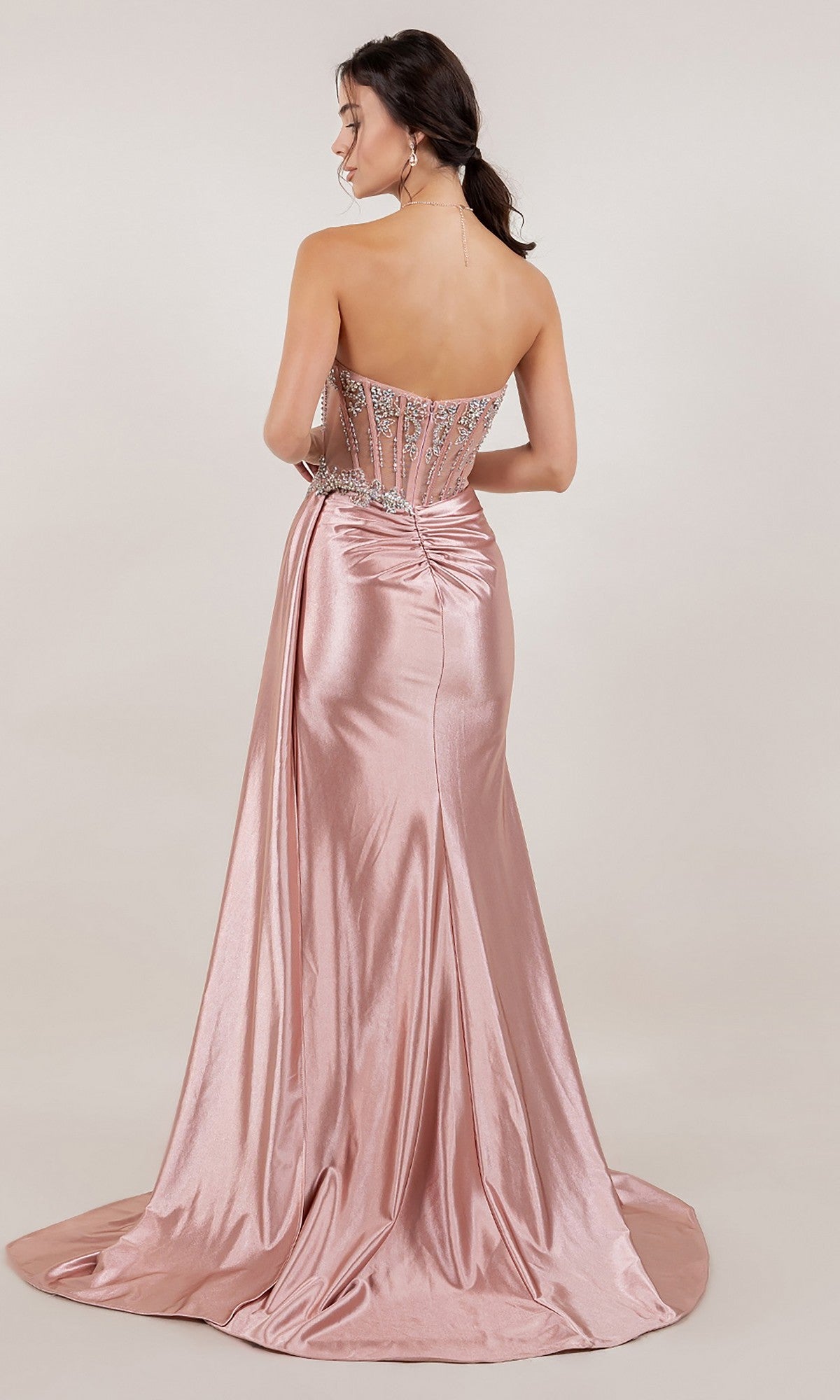 A woman faces away from the camera in the Cinderella Couture 8511J, a strapless metallic pink prom dress with crystal beaded corset bodice and floor-length ruched satin skirt.